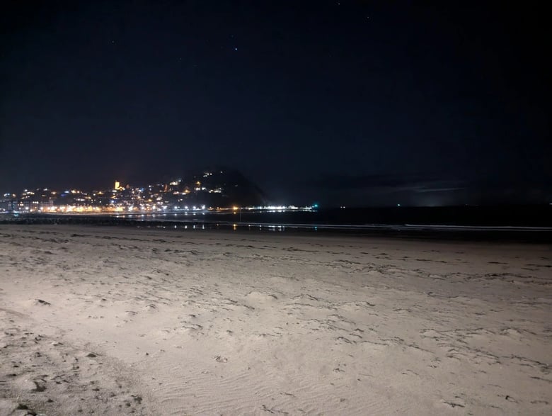 A night-time photo of Minehead beach, showing the headland in the distance. Lights can be seen on the headland, and their reflections in the water. In the foreground, the sand of the beach is illumated.
