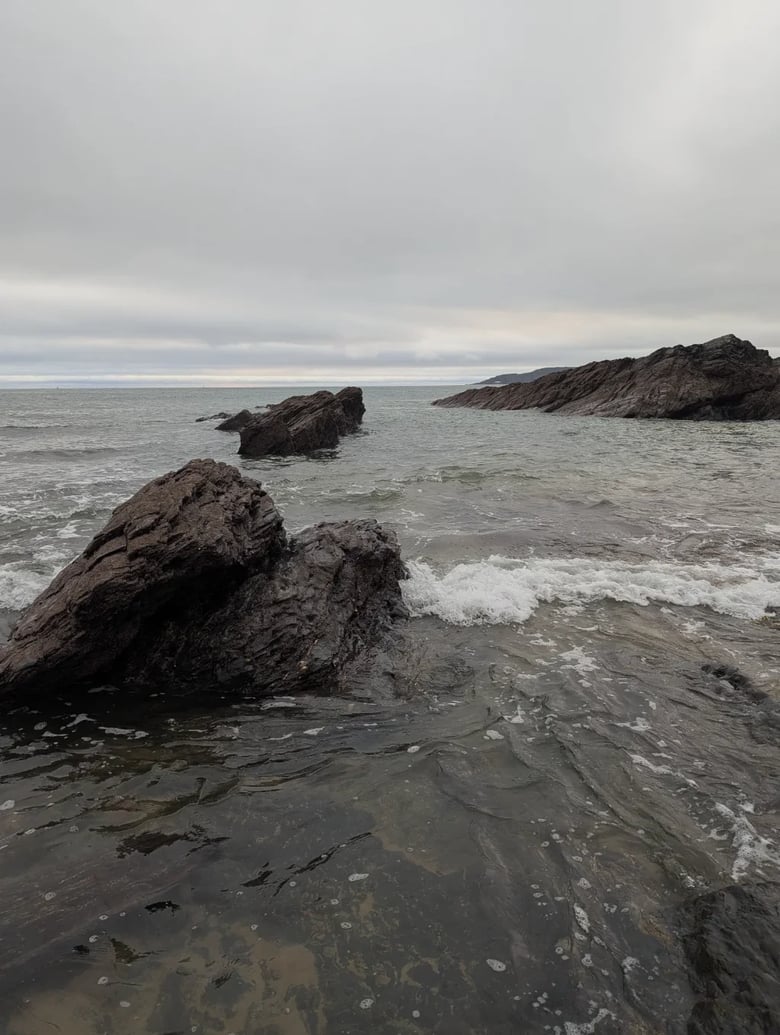 A photo of rocks on a beach, looking out to sea. A small wave has just washed across the lowest rocks. To the left closest to the camera is a rock jutting out of the water. There is a similar rock further back, near the centre of the image, and in the distance a larger rock. The sky is cloudy with a slight hint of sunshine on the horizon.