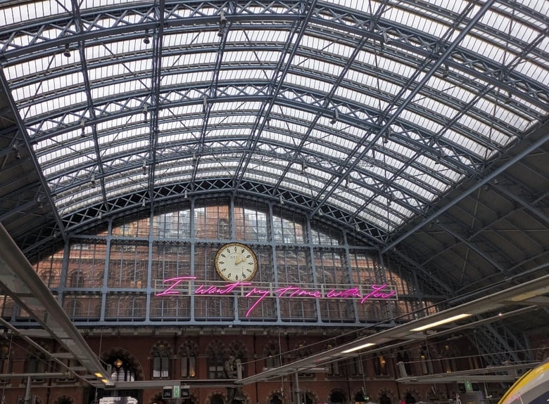 A photo looking at the clock at the St Pancras Eurostar Station. Under the clock face is neon pink writing that says "I want my time with you". At the bottom right of the photo is the yellow and white front edge of a Eurostar train.