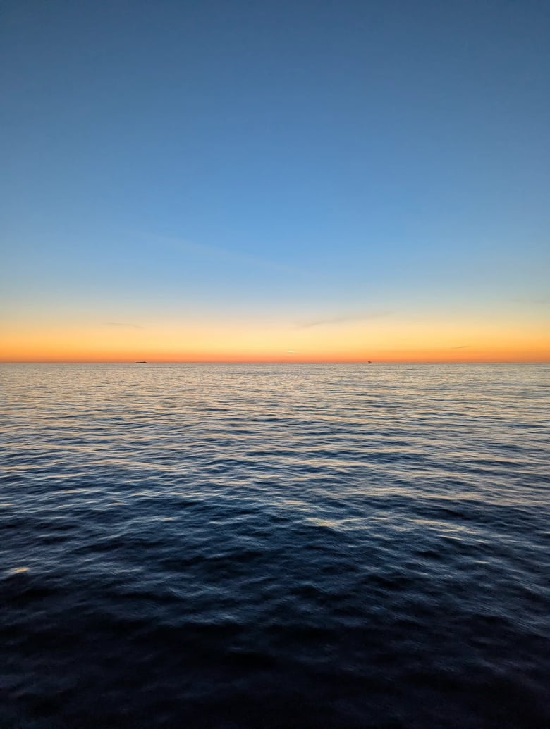 An evening sunset overlooking the North sea. The sky colours graduate from orange through yellow to blue. In the far distance, the silhouette of two ships can be seen on the horizon. The water has small ripples and waves.