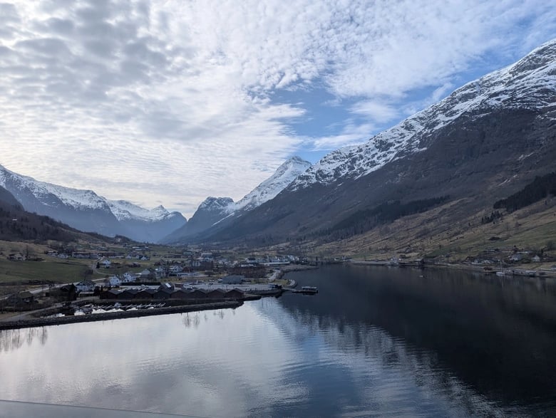 A view of Fjords at Olden, Norway. The sky is bright but cloudy. On either side of the image are steep-sided mountains extending to the horizon. The mountains are capped with snow. At the bottom of the photo is water, and on the left hand side buildings can be seen in the distance on the low ground.