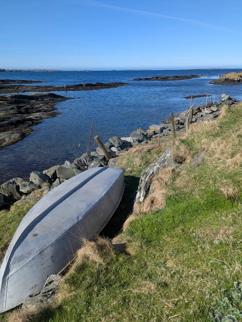 A photo of a rocky shore with a grey boat resting on its side. A blue ocean, a wooden fence, and a distant island create a scenic landscape under clear blue skies.
